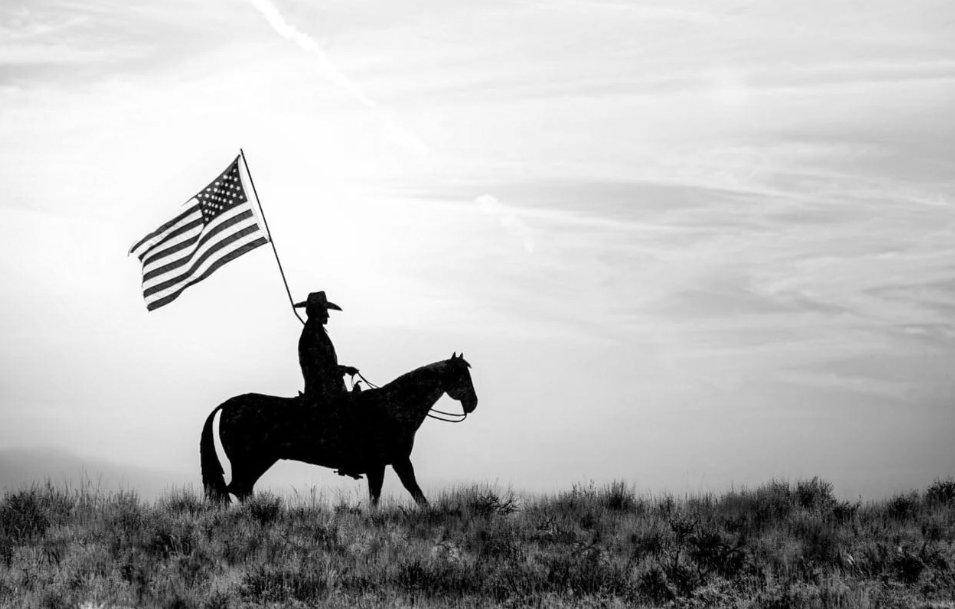 Silhouette of a rider on horseback carrying an American flag across open grassland at dusk